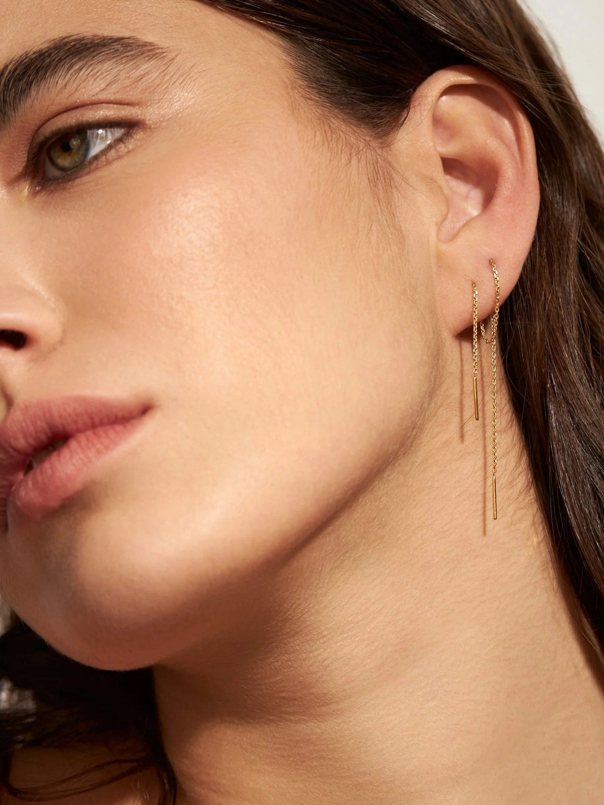 Woman wearing gold ear thread earrings with a thin chain and small bar ends, close-up of the ear