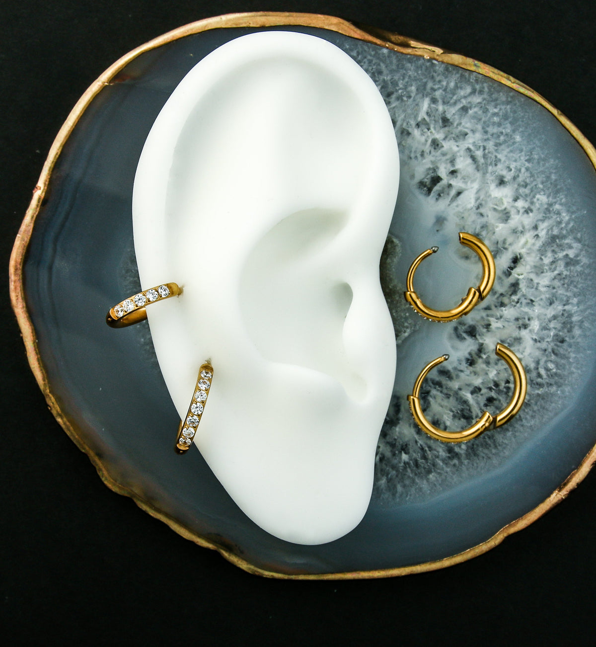 Gold PVD Titanium Clear CZ Hinged Hoop Huggie Earrings, worn on the helix of a display ear with two opened hoops beside it